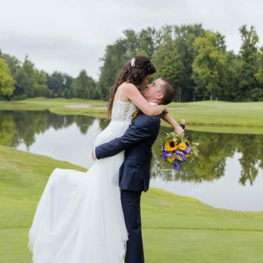 Groom lifts bride holding a bouquet of flowers by pond.