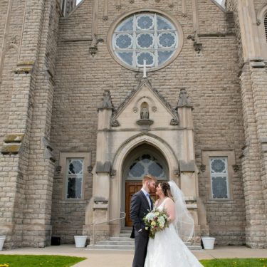A bride and groom standing facing each other in front of a church.
