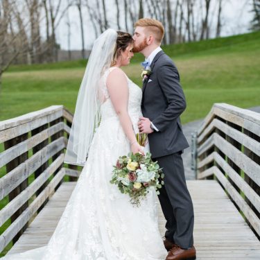 Groom kissing forehead of bride on a wooden bridge in MI